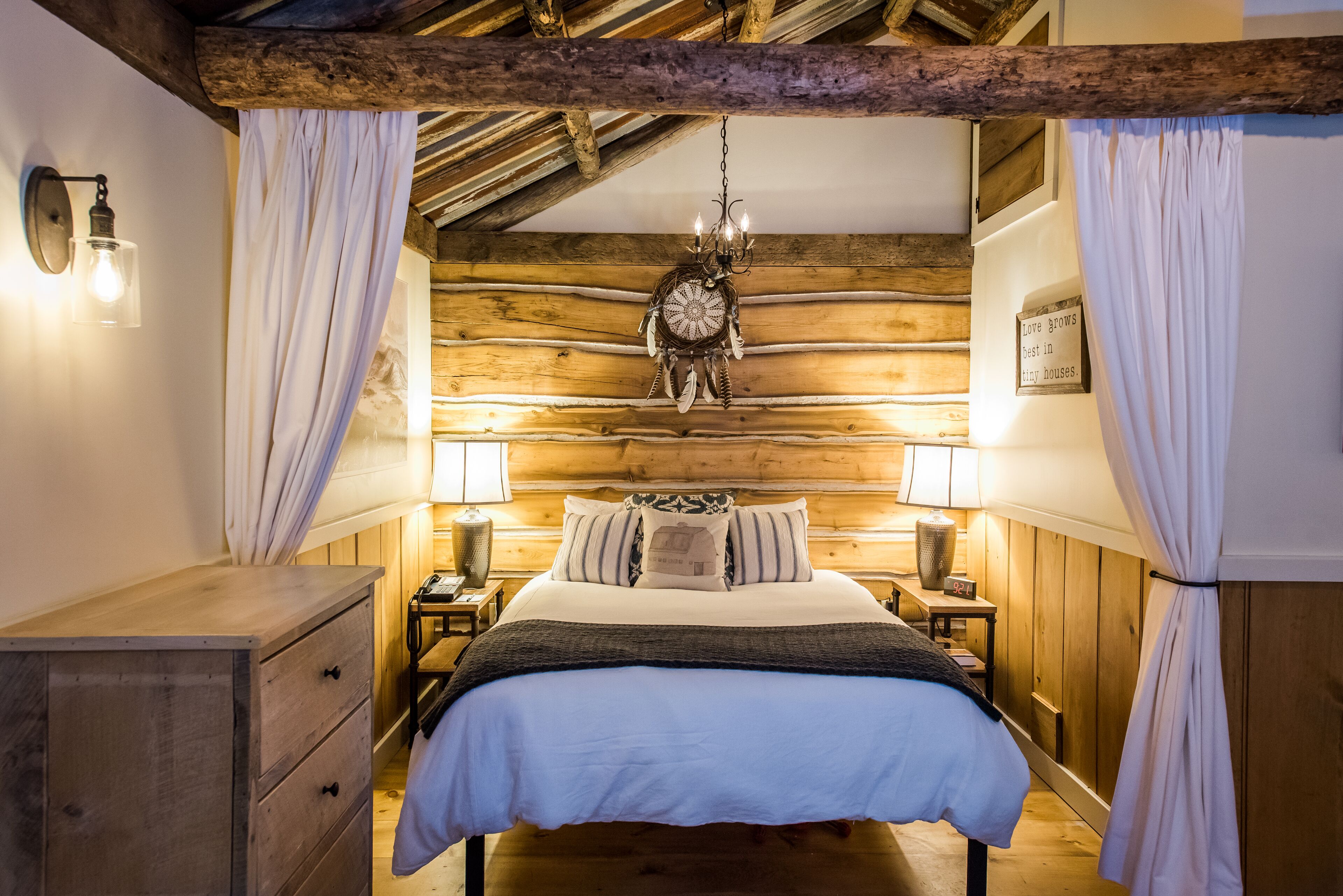 A cozy bedroom with a wooden ceiling, a bed with white bedding, two bedside tables with lamps, and a decorative chandelier.