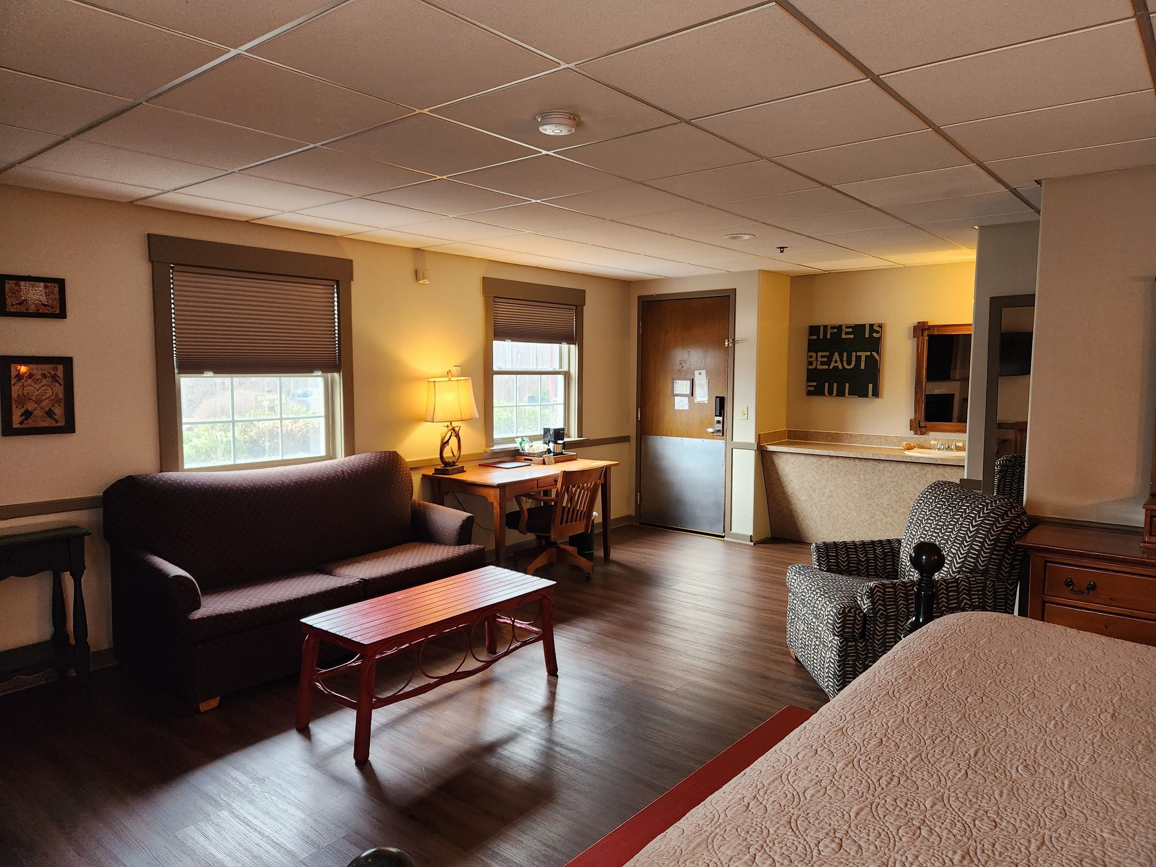 A hotel room with a wooden floor, a sofa, a small table, a desk, and a chair. There is a window with blinds, a lamp, and a mirror on the wall.