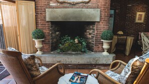 Un espace extérieur accueillant avec des fauteuils en osier et une petite table ronde, une cheminée en briques agrémentée de plantes décoratives et un miroir encadré accroché au mur.