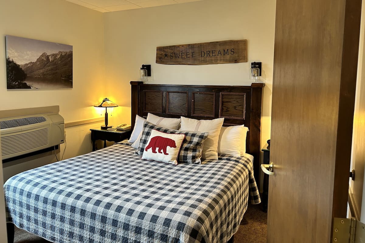 A neatly made bed with a checkered bedspread, a mounted picture of a mountain landscape, and a wooden headboard with a sign above it.