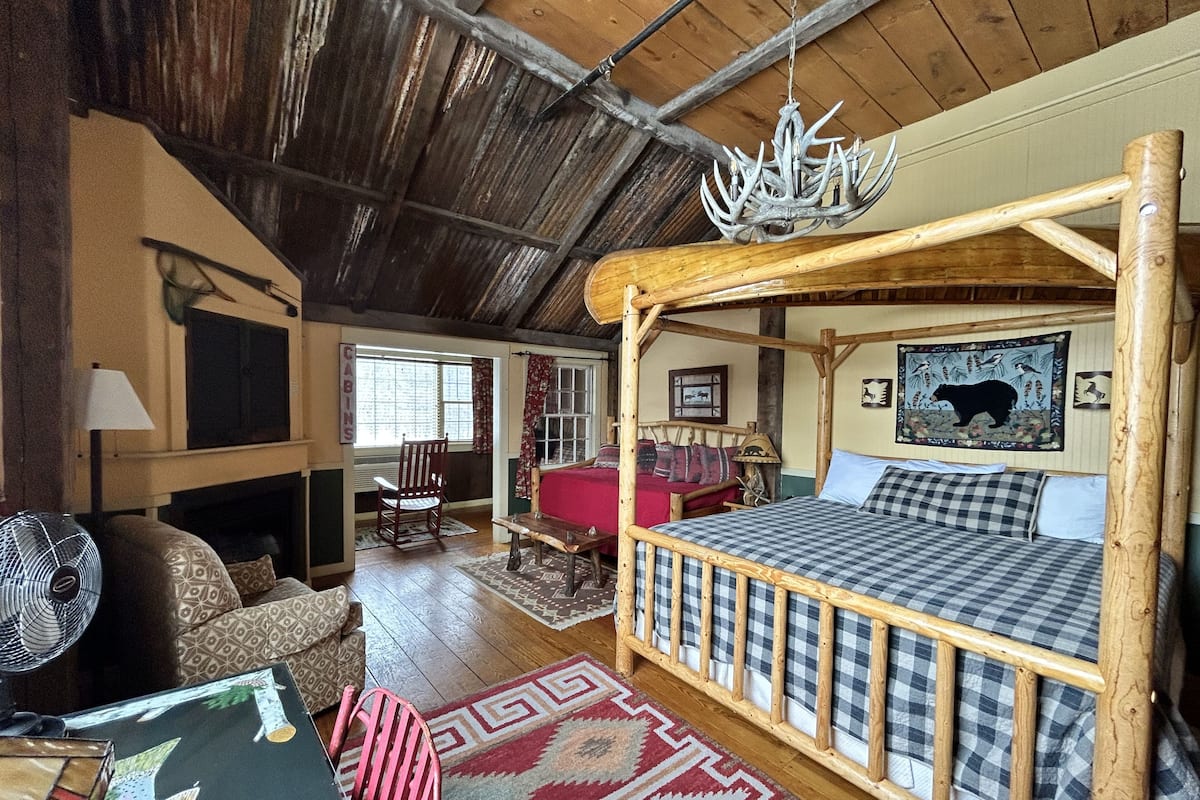 A rustic cabin interior with a wooden bed, a rocking chair, a fireplace, and a chandelier.