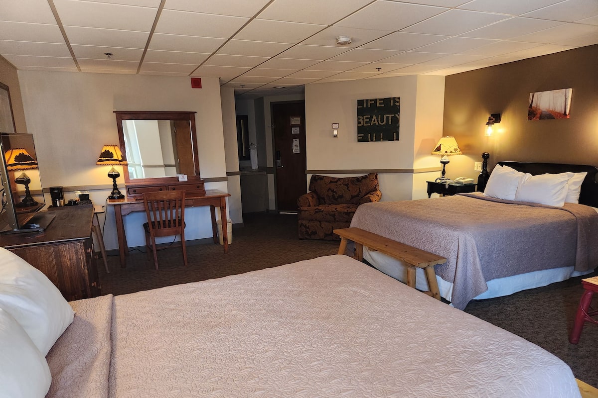 A hotel room with two beds, a desk, a chair, a bench, and a small kitchenette.