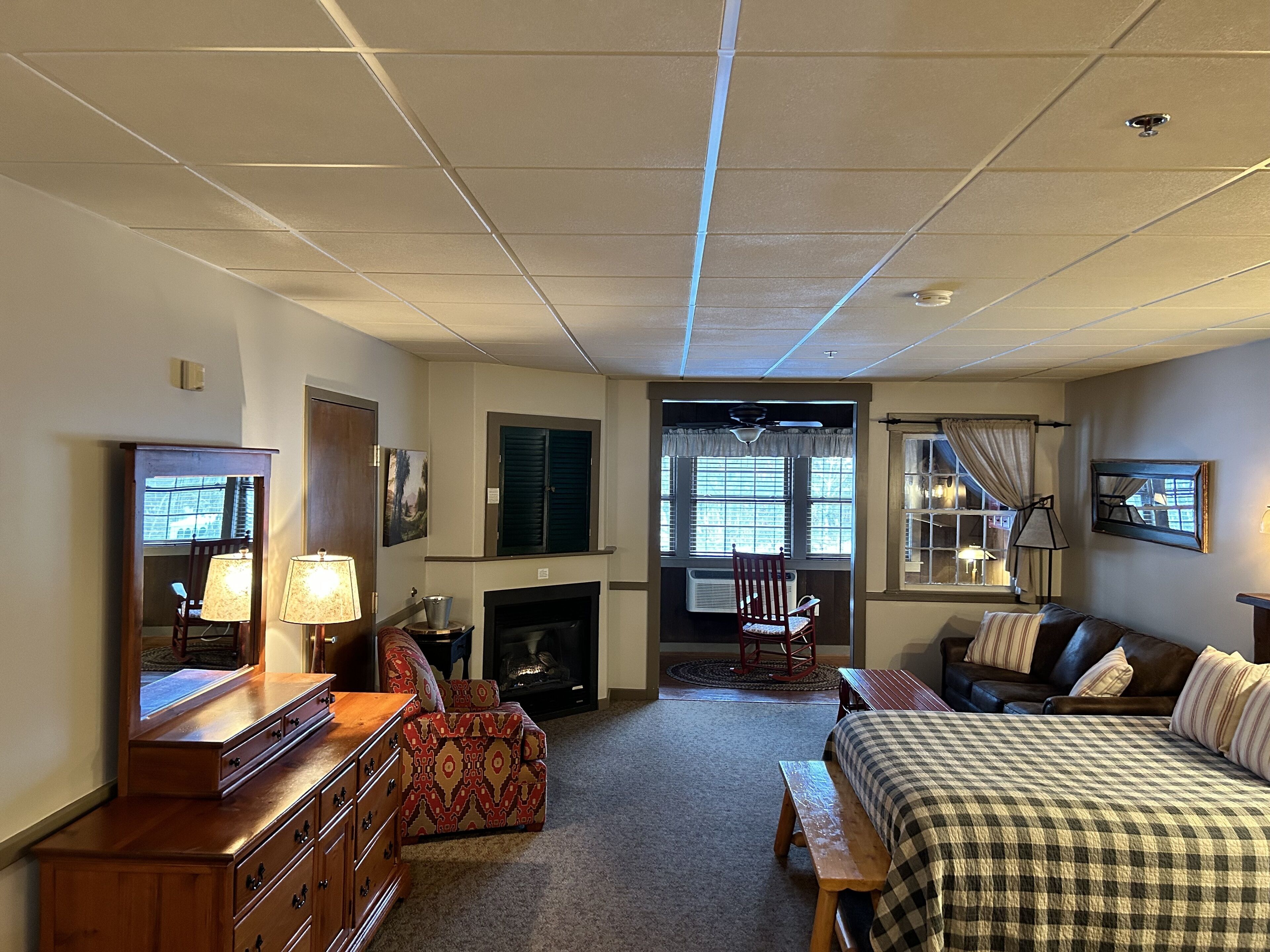 A hotel room with a fireplace, a bed, a wooden dresser, a chair, and a window with curtains.