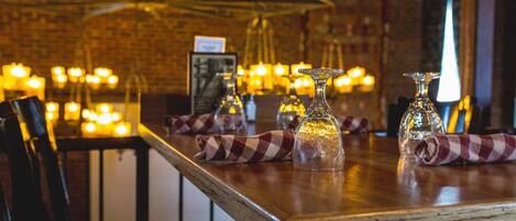 A restaurant interior with a rustic brick wall, wooden tables, and hanging light fixtures.