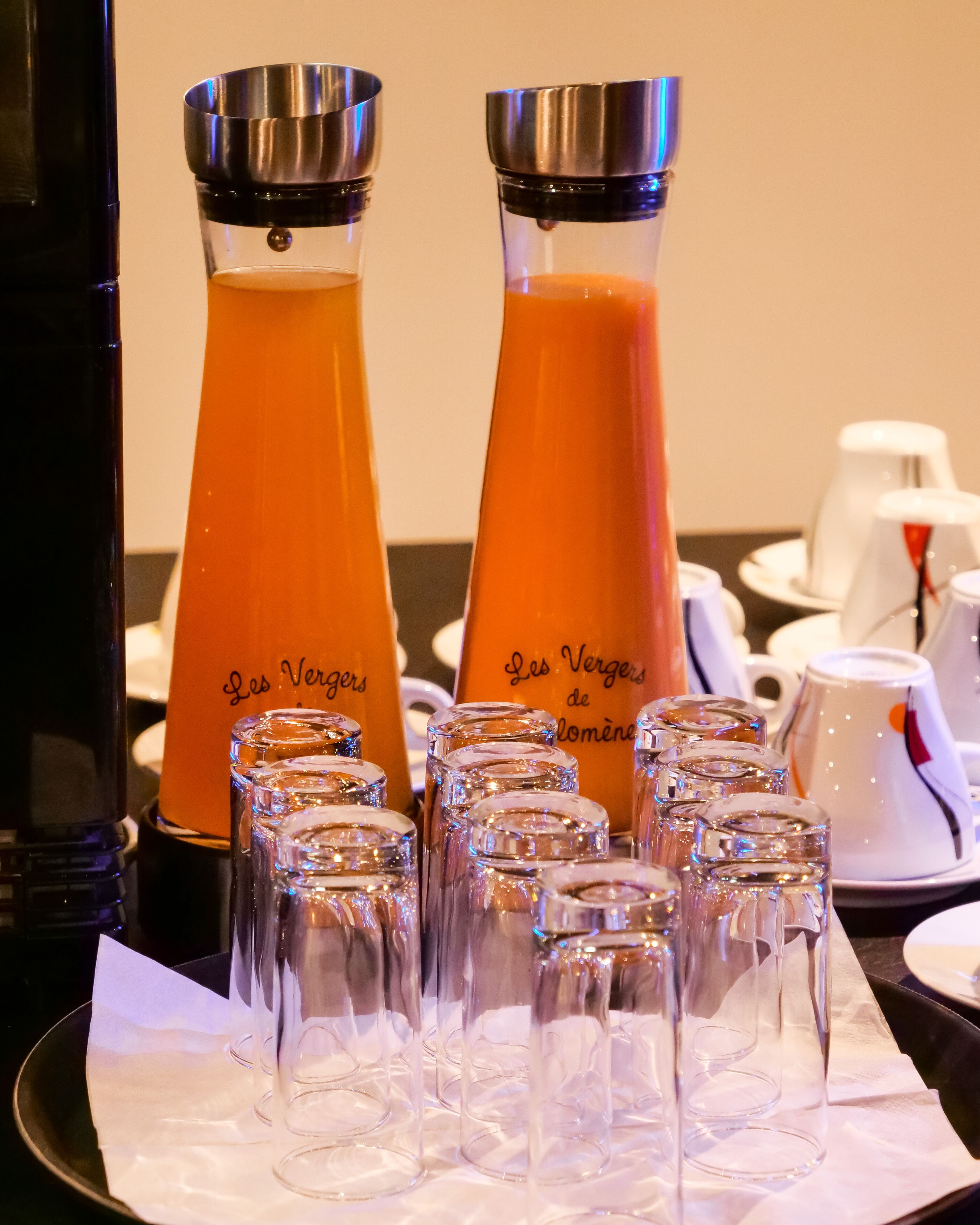 Two orange juice dispensers labeled "Les Vergers de Lomène" and several clear glasses arranged on a tray.