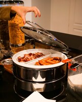 A buffet setup with a large pot containing sausages and bacon, a hand reaching for the lid, and a container of cereal in the background.