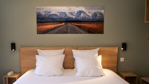A hotel room with a large bed, two bedside tables, a mounted painting of a mountain road, and a wall-mounted phone. - Motel 991 (Viviers-du-Lac)