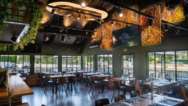 A restaurant interior with multiple dining tables and chairs, large windows offering views of the outside, and hanging plants. - Motel 991 (Viviers-du-Lac)