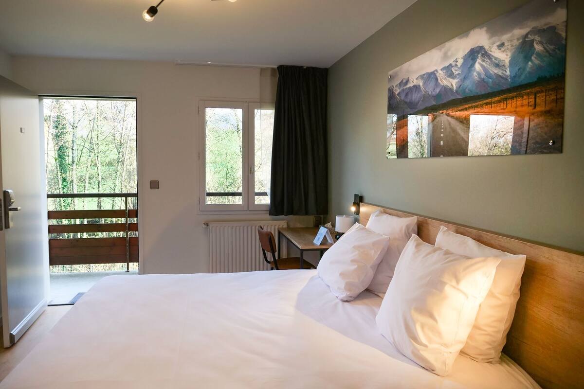 Ein Hotelzimmer mit einem großen Bett, einem Schreibtisch und Blick auf einen Wald durch ein Fenster.
