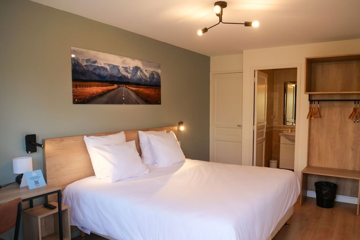 A hotel room with a bed, a nightstand, a desk, a coat rack, and a painting on the wall.