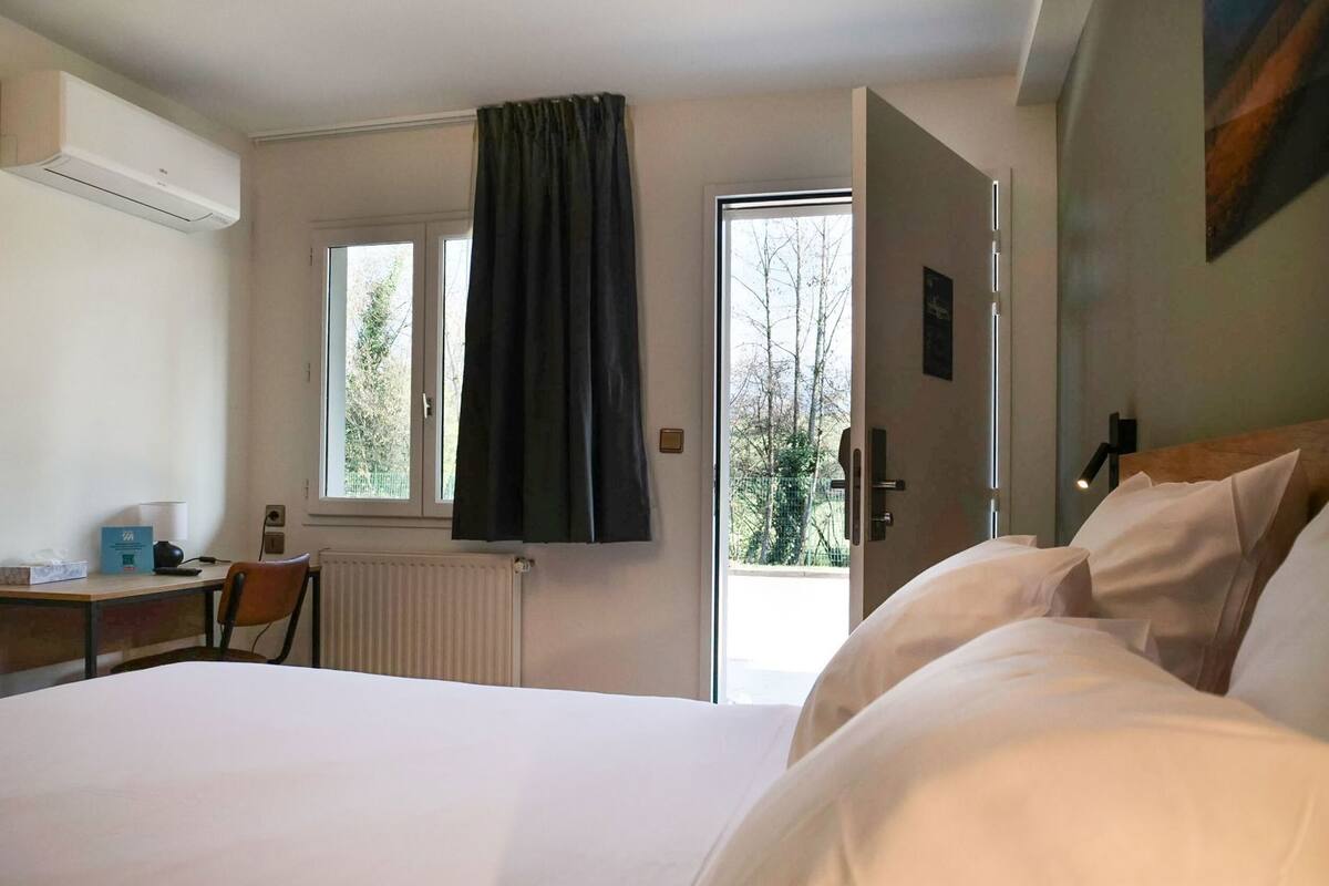 Ein Hotelzimmer mit Bett, Schreibtisch, Stuhl, Heizkörper, einem Fenster mit Vorhängen und einer Tür.