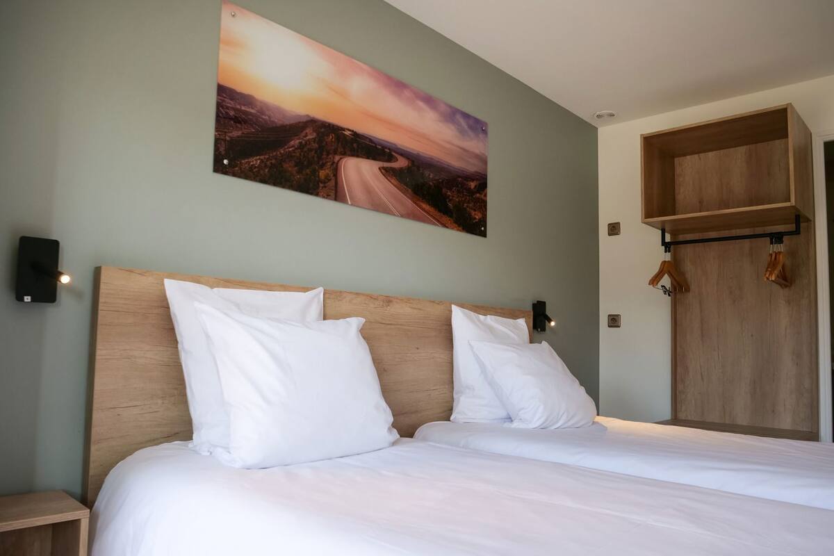 Ein Schlafzimmer mit einem hölzernen Kopfteil, weißer Bettwäsche, einem an der Wand hängenden Landschaftsbild und einem Holzschrank mit Kleiderstange.