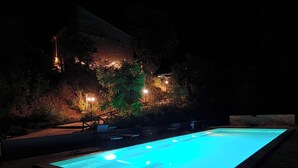 Una piscina exterior de noche, rodeada de árboles y un muro de piedra.