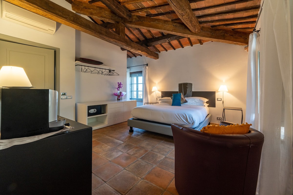 Ein Schlafzimmer mit einem Bett, Nachttischlampen, einem Schreibtisch mit Lampe, einem Sessel und einem Fenster mit Vorhängen.