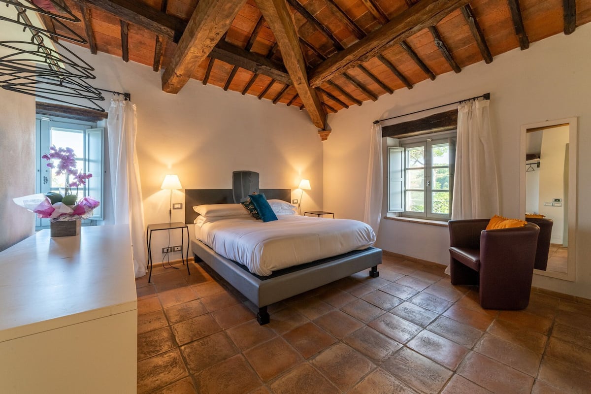Ein Schlafzimmer mit einem Bett, zwei Nachttischlampen, einem Fenster, einem Sessel und einem kleinen Tisch mit Lampe.