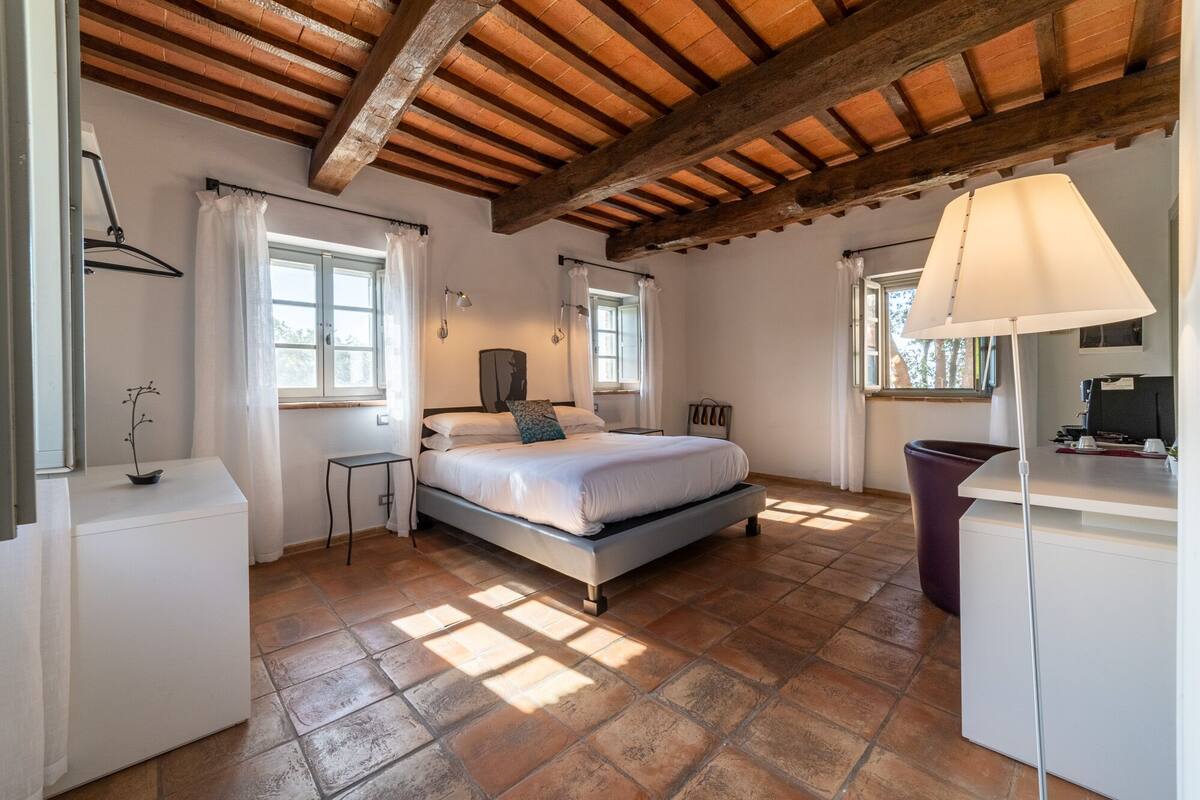 Ein Schlafzimmer mit Bett, Nachttischen, Schreibtisch und Stuhl. Es gibt Fenster mit Vorhängen, eine Decke mit Holzbalken und eine Lampe auf dem Schreibtisch.