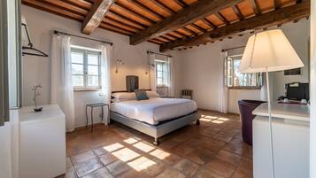 Ein Schlafzimmer mit Bett, Nachttischen, Schreibtisch und Stuhl. Es gibt Fenster mit Vorhängen, eine Decke mit Holzbalken und eine Lampe auf dem Schreibtisch.