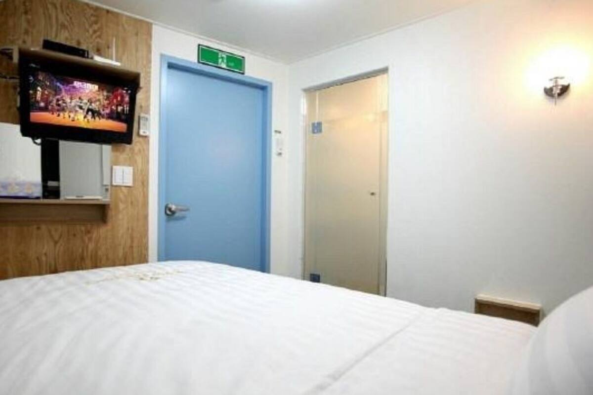 A hotel room with a bed, a television mounted on the wall, a blue door, and a light fixture on the wall.