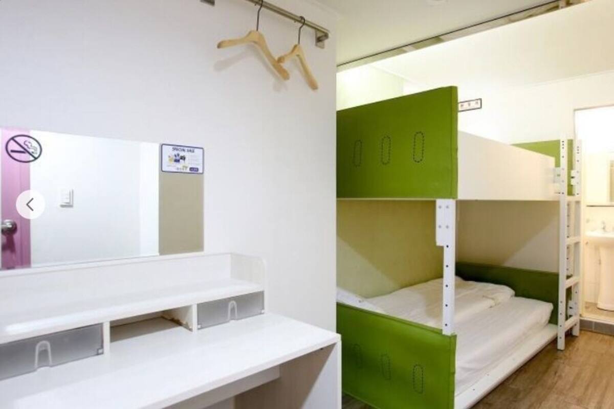 A dormitory room with bunk beds, a white desk, a mirror, and a bathroom with a sink and toilet.