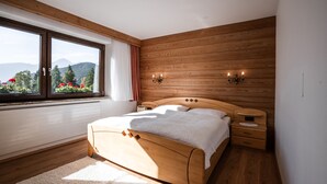 A bedroom with a wooden bed, a nightstand, a wall-mounted lamp, and a window with a view of mountains and flowers. - bright apartment with a large sun terrace and access to the garden (Reith bei Seefeld)