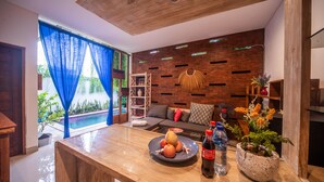 Un salon moderne avec un plafond en bois, un mur décoré d’un motif en briques, un canapé avec des coussins, une table basse avec des fruits et des boissons, et une vue sur une piscine à travers des rideaux bleus.