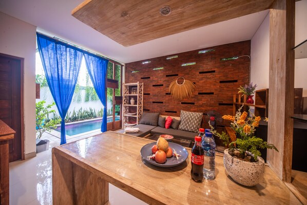 A modern living room with a wooden ceiling, a brick-patterned wall, a sofa with cushions, a coffee table with fruit and drinks, and a view of a swimming pool through blue curtains.