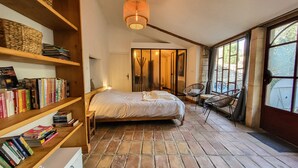 Een knus slaapkamer met een groot bed, een boekenplank vol boeken, een rieten stoel en uitzicht door een raam.