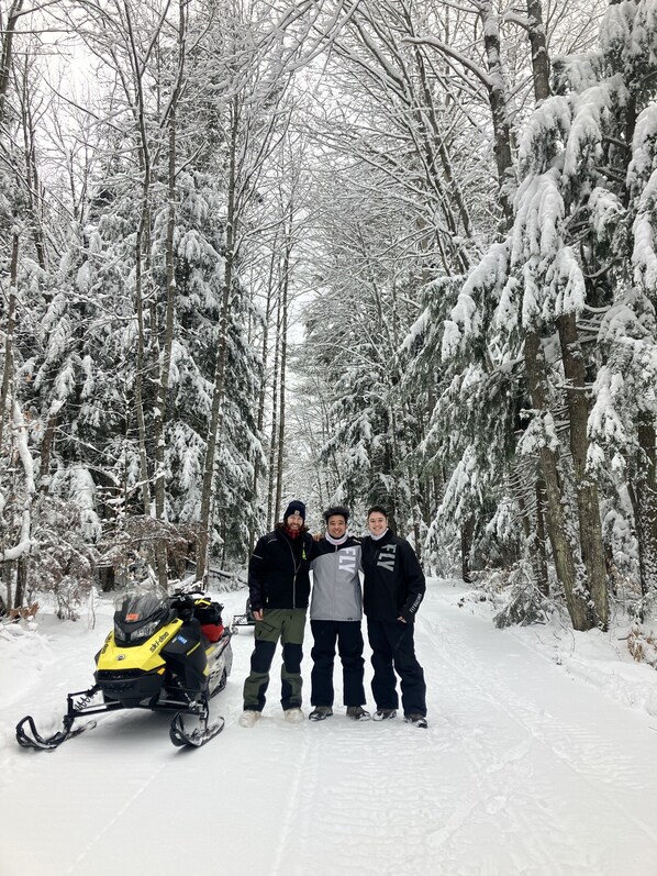 Three people standing in a snowy forest with a snowmobile parked nearby.