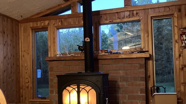 A cozy room with a wood-burning stove, large windows, and a fireplace mantel. - Cozy Cabin located directly on the ORV trail. Peace & seclusion. (Hulbert)