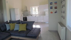 A modern living room with a grey sectional sofa, a white radiator, and a kitchen in the background. - Beautiful 3-bed House in Chania With Garden (Platanias)