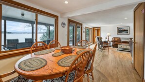 A spacious dining area with a large wooden table, multiple chairs, and a view of a lake through large windows. - Waterfront home sleeping 10 located on Cummings Cove on Lake Winnipesaukee (Meredith)