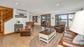 A spacious living room with a wooden staircase, a fireplace, and a view of a lake. - Waterfront home sleeping 10 located on Cummings Cove on Lake Winnipesaukee (Meredith)