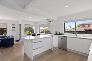 A modern kitchen with white cabinetry, a central island, and a dishwasher. There is a dining area with a blue sofa and a large window offering a view of the neighborhood. - Mount Maunganui Getaway: Waitui Wonder House (Mount Maunganui)