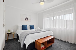 A bedroom with a bed, a small side table, a ceiling fan, and a window with sheer curtains. - Mount Maunganui Getaway: Waitui Wonder House (Mount Maunganui)