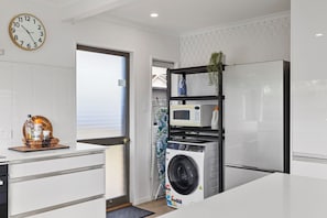 A modern kitchen with a built-in washing machine, a microwave, and a clock on the wall. - Mount Maunganui Getaway: Waitui Wonder House (Mount Maunganui)