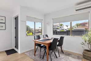 A modern room with a dining table, four chairs, a black rug, and large windows offering a view of the neighborhood. - Mount Maunganui Getaway: Waitui Wonder House (Mount Maunganui)