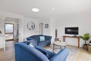 A modern living room with a sofa, a coffee table, a TV, and a small side table with decorative items. - Mount Maunganui Getaway: Waitui Wonder House (Mount Maunganui)