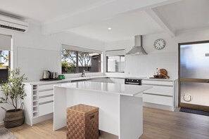 A modern kitchen with white cabinetry, a large island, a built-in oven, and a window overlooking a backyard. - Mount Maunganui Getaway: Waitui Wonder House (Mount Maunganui)
