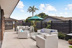 An outdoor patio area with wicker furniture, a green umbrella, and a view of neighboring houses and trees. - Mount Maunganui Getaway: Waitui Wonder House (Mount Maunganui)