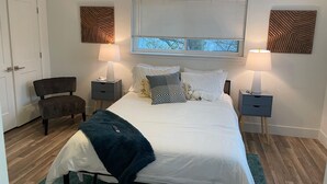 A bedroom with a bed, two bedside tables, a chair, a window with blinds, and decorative wall panels. - Casa Wooten -Contemporary Condo (Austin)