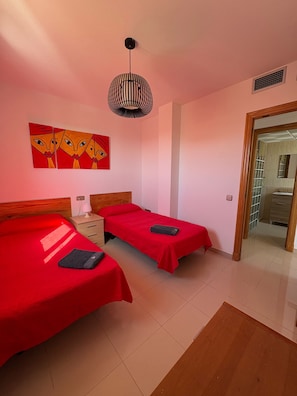 A room with two beds, a wooden headboard, a bedside table with a lamp, and a wall-mounted artwork.