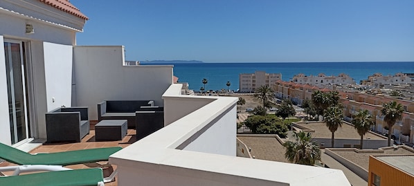 A balcony with outdoor furniture, a clear view of the sea, and a residential area with palm trees.