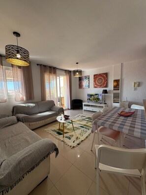 A living room with a sofa, a coffee table, and a TV. There is a dining table with chairs. The room has a large window with curtains.