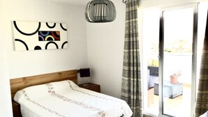 Une chambre avec un lit en bois, une table de chevet avec une lampe, un luminaire suspendu et une vue sur l’extérieur grâce à une porte-fenêtre coulissante.