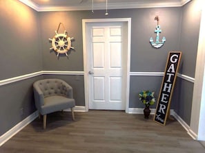 A hallway with a white door, a tufted armchair, a wall-mounted anchor decoration, and a 'GATHER HERE' sign.