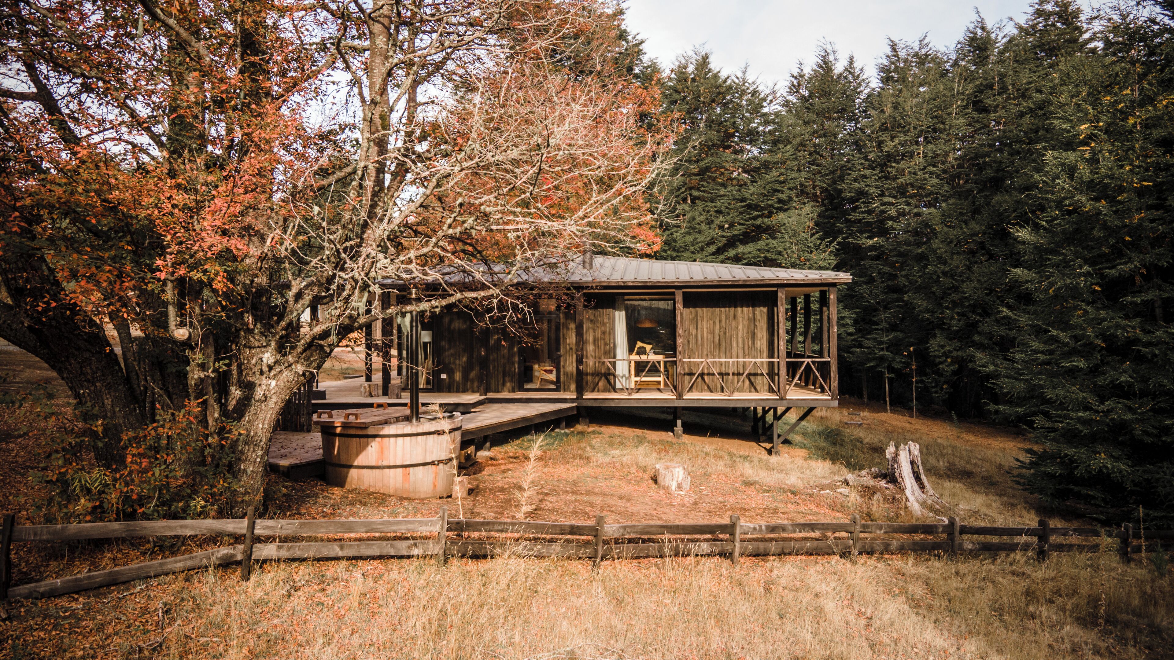 A rustic cabin with a wooden deck and a large barrel, surrounded by trees and a wooden fence.