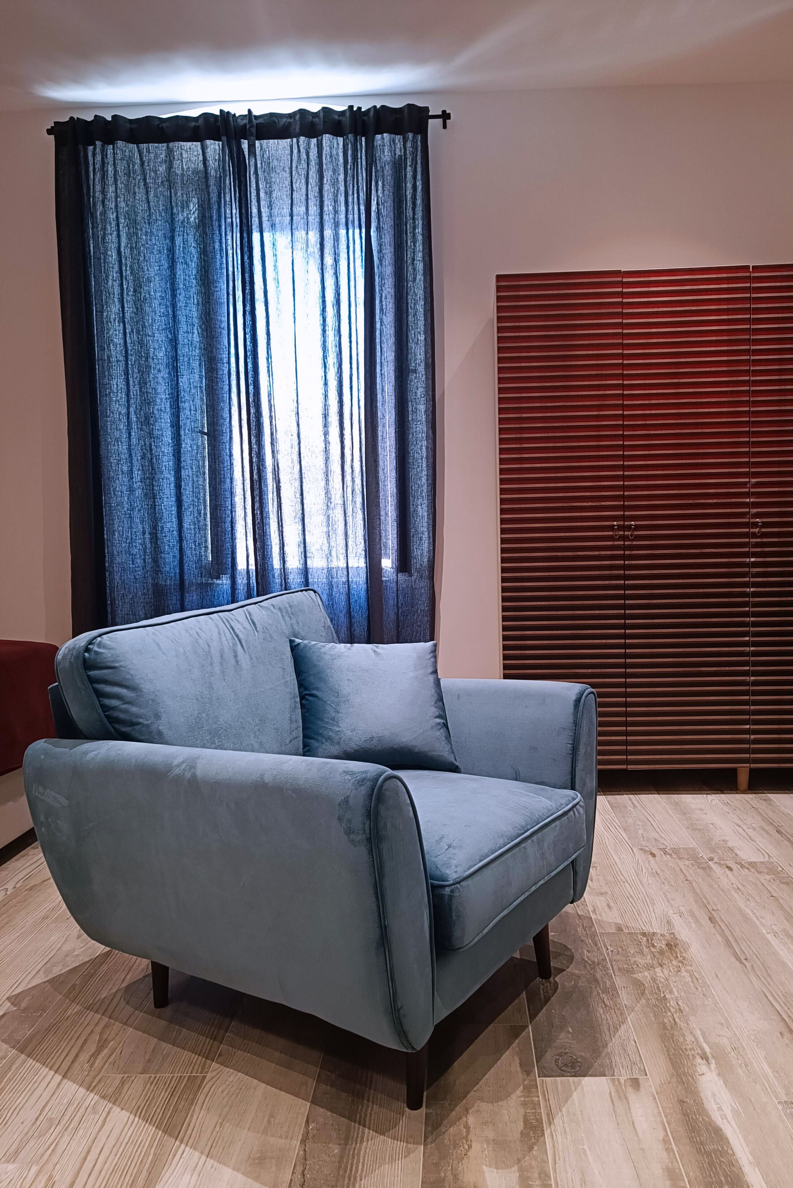 A room with a blue sofa, a window with sheer curtains, and a wooden wardrobe.