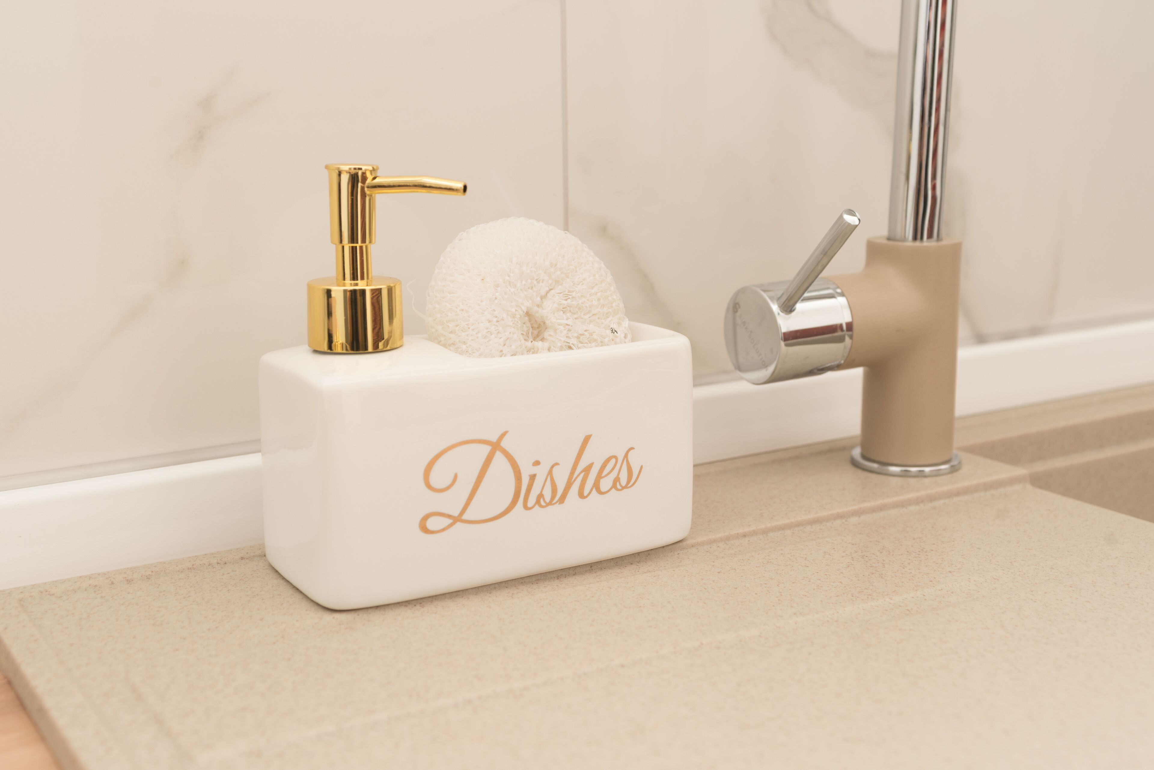 Un lavamanos de baño con un grifo dorado, un dispensador de jabón color beige y un recipiente blanco para lavar trastes con la etiqueta 'Dishes'.