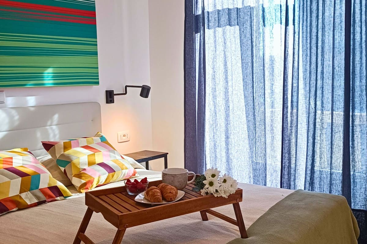 A hotel room with a bed, a bedside table, a tray with pastries, a colorful wall hanging, and a window with curtains.