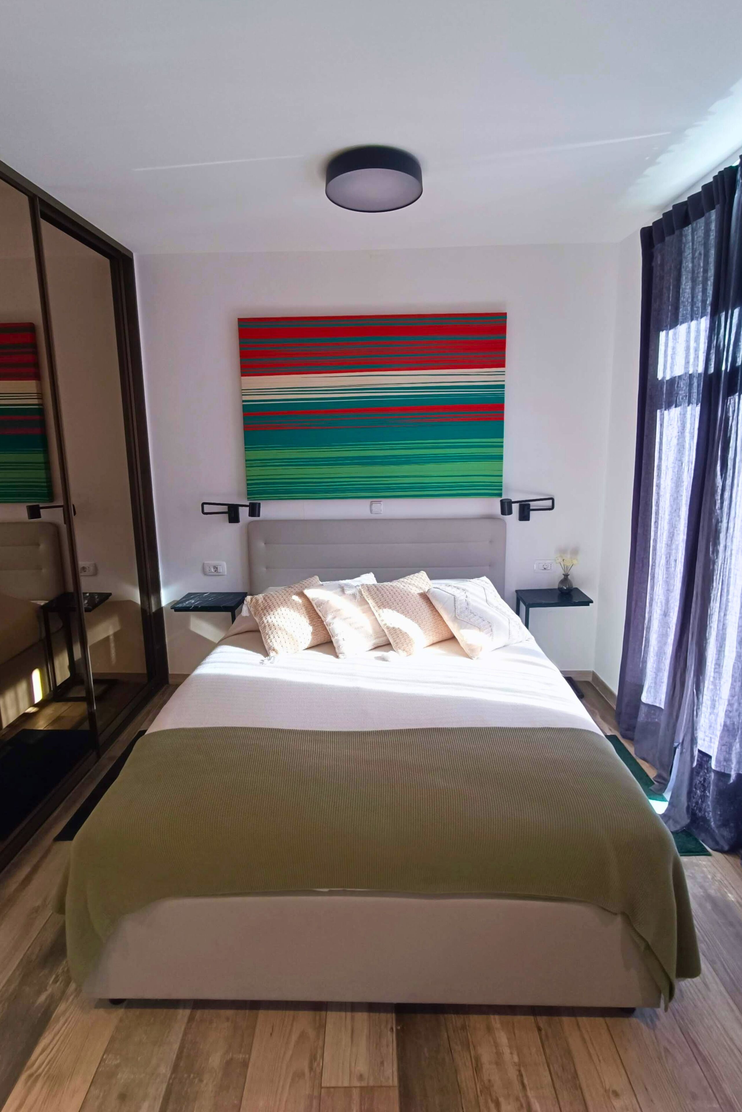 Habitación de hotel moderna con una cama grande, un espejo, una mesita de noche con lámpara y un cuadro abstracto y colorido en la pared.